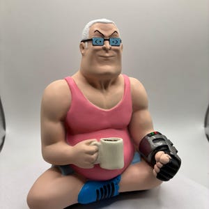 Peut inclure: Figurine d'un homme musclé aux cheveux blancs et lunettes, vêtu d'un débardeur rose et d'un short bleu, tenant une tasse à café. Il porte un appareil noir et gris au poignet. La figurine est assise en tailleur.