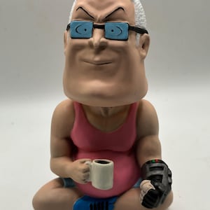 Peut inclure: Figurine d'une personne assise avec une expression sévère, portant des lunettes bleues et un débardeur rose. La figurine tient une tasse blanche dans une main et un appareil noir dans l'autre. La figurine est assise, les jambes croisées, et porte des chaussures bleues.