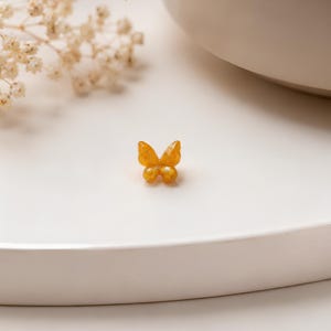 Puede incluir: Un pequeño dije en forma de mariposa de color ámbar con un acabado brillante y translúcido. La mariposa presenta alas redondeadas y una superficie lisa y pulida, captando la luz para resaltar su cálido tono dorado anaranjado.