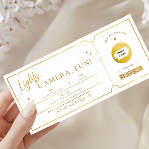 May include: A white rectangular event ticket with gold foil borders and text. It features the words Lights, CAMERA, FUN!, a gold circular scratch-off area, a barcode, and fields for date, time, and location. The design includes small star accents.