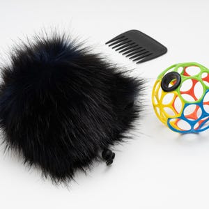Puede incluir: Una cubierta de micrófono negra y peluda con un cordón, un peine negro y un juguete de pelota de plástico de colores con agujeros. La pelota es principalmente amarilla, verde, roja y azul.
