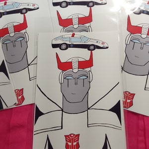 Puede incluir: Un coche de policía de dibujos animados blanco y negro con una cabeza de robot roja y blanca con un símbolo de Autobot rojo en el pecho. El robot tiene un casco blanco con cuernos rojos y una cara blanca con ojos negros.