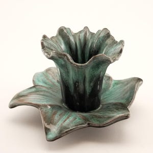 May include: A ceramic teal-coloured flower-shaped candle holder. The candle holder has a dark teal interior and a matching saucer base. The edges of the flower petals are slightly ruffled, adding a unique touch to the design.