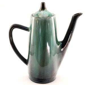 May include: A vintage ceramic coffee pot with a tall, slender design. The pot has a gradient of green hues, from light at the top to a darker shade at the base. The handle and spout are black. The lid has a small, dark knob.