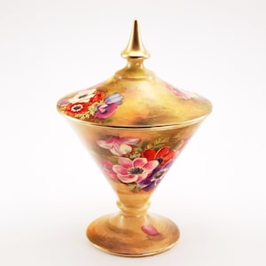 May include: A gold-coloured, lidded decorative jar with a conical body and a pedestal base. The jar features hand-painted floral designs in red, pink, and purple. The lid is topped with a gold finial. The jar is likely made of ceramic or porcelain.