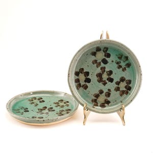 Peut inclure: Deux assiettes en céramique avec un glaçage turquoise et un motif floral. Les assiettes ont un design moucheté avec des accents marron et blancs. Une assiette est présentée sur un support doré. Les assiettes sont faites à la main.