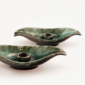 May include: Two ceramic candle holders in a leaf shape. The holders are dark green with a glossy finish and a dark brown base. Each has a circular opening for a candle.