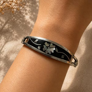 Peut inclure: Un bracelet manchette argenté avec une incrustation noire et un motif floral. Le bracelet présente des incrustations de fleurs et de feuilles en nacre. Le bracelet est porté au poignet.