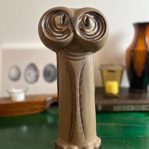 May include: A tan ceramic owl figurine with large, detailed eyes and a textured body. The owl has a long, slender body with a decorative pattern down the front. The figurine is standing on a green surface.