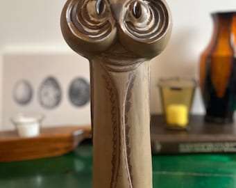 Könnte beinhalten: Eine braune Keramik-Eulenfigur mit großen, detaillierten Augen und einem strukturierten Körper. Die Eule hat einen langen, schlanken Körper mit einem dekorativen Muster auf der Vorderseite. Die Figur steht auf einer grünen Oberfläche.