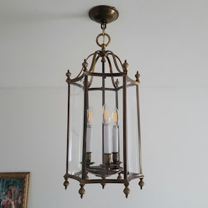 Peut inclure: Un luminaire suspendu hexagonal en laiton avec des panneaux de verre transparents. L'intérieur comprend trois fausses bougies. Le luminaire est suspendu au plafond par une chaîne et un auvent circulaire. Des finials décoratifs ornent le haut et le bas.