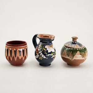 May include: Three pieces of hand-painted pottery. A small brown cup with an orange and white design, a dark blue jug with floral accents, and a round sugar bowl with a lid featuring a green and brown forest design.