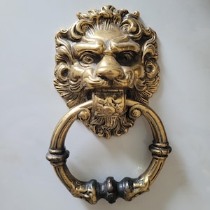 Op de afbeelding: Een messing deurklopper met een leeuwenkopontwerp. De leeuw heeft een gedetailleerde manen en een ringhandvat. De klopper is gemonteerd op een witte ondergrond.