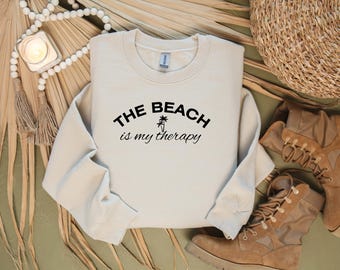 以下が含まれることがあります： ベージュのトレーナーに、黒い文字で「THE BEACH is my therapy」と書いてあります。テキストはアーチ状になっており、ヤシの木のグラフィックが含まれています。