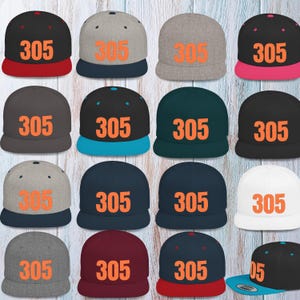 May include: A collection of flat-brimmed baseball caps in various colors, including black, gray, navy, and white. Each cap features the number "305" in orange block letters on the front. The brims are in contrasting colors.