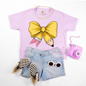 May include: A light purple t-shirt with a large yellow bow and pencil graphic, paired with denim shorts, checkered sneakers, sunglasses, a beaded necklace, and a pink camera. The outfit is styled on a white background.