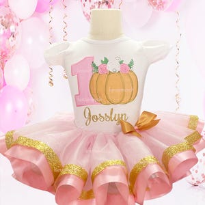May include: A pink tulle tutu with gold glitter trim and a white shirt with a pink number one, a pumpkin, and the name "Josslyn" embroidered on it.