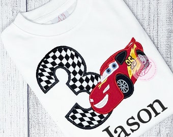 Puede incluir: Camiseta blanca con un coche rojo Rayo McQueen y un número 3 a cuadros blanco y negro. El nombre Jason está escrito debajo del coche.