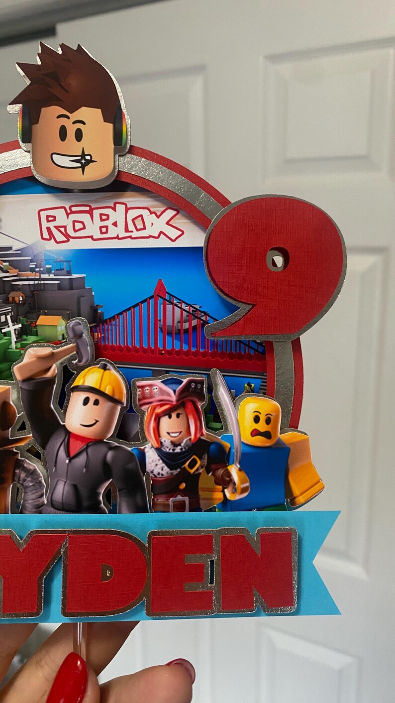 May include: A red and silver Roblox themed cake topper with a number 9, a Roblox character, and the name "RYDEN".