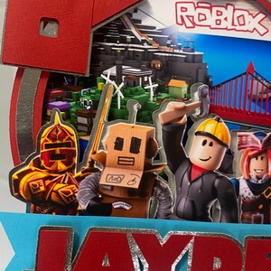 May include: A red and silver Roblox themed cake topper with a Roblox logo, a character with headphones, and a name that reads "JAYDE".