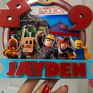 May include: A red and silver cake topper with a Roblox logo, a Roblox character, and the name Jayden. The cake topper features a red and blue background with a bridge and buildings.
