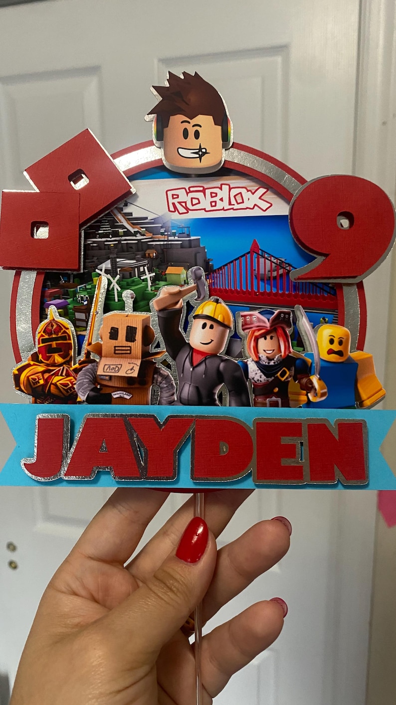 May include: A red and silver cake topper with a Roblox logo, a Roblox character, and the name Jayden. The cake topper features a red and blue background with a bridge and buildings.