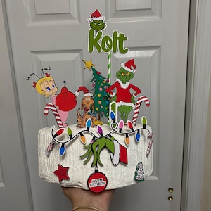 Puede incluir: Un pastel blanco decorado con un tema de Grinch. El pastel tiene luces verdes y rojas, una mano verde de Grinch, un adorno rojo, un pequeño árbol de Navidad y un letrero rojo que dice "Feliz cumpleaños". El pastel también tiene un personaje verde de Grinch, un personaje rojo y blanco de Cindy Lou Who, un personaje marrón de Max the Dog y un árbol de Navidad verde.