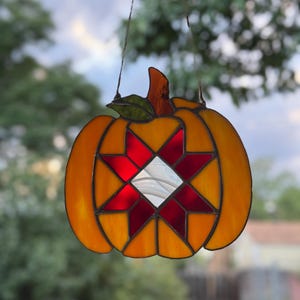 May include: Stained glass pumpkin suncatcher with an orange body, a green leaf, and a brown stem. The pumpkin features a star design with red and white glass. The suncatcher is hanging from a thin wire.