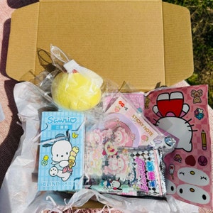 May include: A pink and white box filled with various items, including a yellow sponge ball, a blue and white blind box with the text "Sanrio" and "Blind Box", a pink and white sticker sheet with the text "Sticky Note", and a blue and white package with Japanese text.