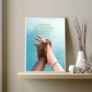 May include: A framed print with a quote by Nelson Mandela: "A winner is a dreamer who never gives up". The print features a photo of two hands clasped together, one black and one white, against a blue sky background.