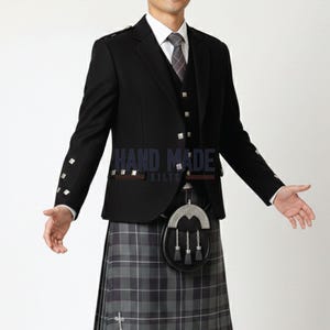 May include: A black wool jacket with silver buttons, a matching waistcoat, and a grey plaid kilt. The kilt has a traditional pattern in grey and black. A sporran hangs from the waist. The text "HAND MADE KILTS" is visible.