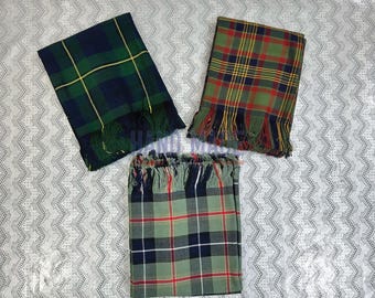 May include: Three folded tartan scarves in shades of green, blue, red, and yellow. Each scarf has a fringed edge. The scarves are arranged on a patterned, light gray surface.
