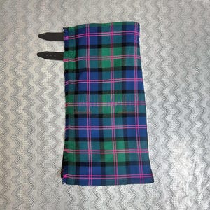 Könnte beinhalten: Ein gefalteter Kilt in einem blau, grün, schwarz und rosa karierten Muster. Der Kilt hat zwei schwarze Lederriemen mit Schnallen. Der Stoff hat einen Fransenrand.