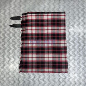 May include: A rectangular piece of fabric with a black, red, and white plaid pattern. Two black leather straps are attached to the top edge. The fabric has a fringed edge. The check design is a traditional pattern.