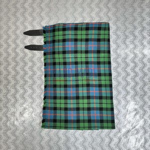 May include: A folded green, blue, and black plaid kilt with two black leather straps. The kilt has a fringed edge. The plaid pattern includes orange stripes. The kilt is made of a woven fabric.