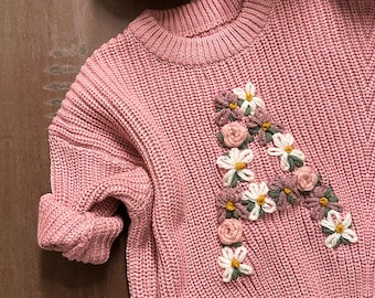 Puede incluir: Un suéter de punto rosa con una letra A bordada con flores en la parte delantera.