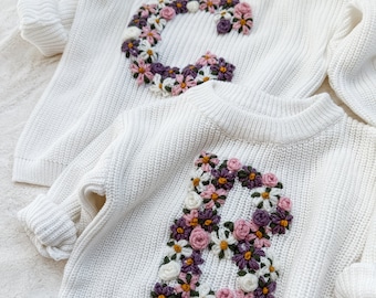 Peut inclure: Deux pulls en tricot blanc avec des lettres brodées de fleurs. L'un présente un grand "C" et l'autre un "B", tous deux ornés de fleurs roses, violettes et blanches. Les pulls ont des manches longues.