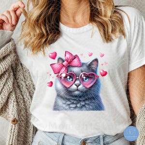 May include: A white t-shirt with a gray cat wearing pink heart-shaped sunglasses and a pink bow with white polka dots. The cat is surrounded by pink hearts.
