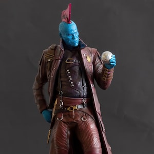 May include: A blue-skinned action figure of a character wearing a red and brown costume with a mohawk. The figure is holding a silver orb in its right hand. The figure is standing on a black base.