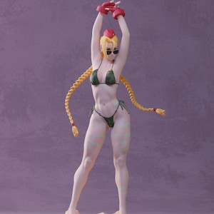 以下が含まれることがあります： 緑色の迷彩柄のビキニトップとボトムを着用した女性のアクションフィギュア。長い金色の三つ編み、赤いボクシンググローブ、サングラスをかけています。フィギュアは砂の台座の上に立っています。