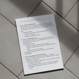 May include: A white sheet of paper with text about decluttering tips, including small wins, tackling bigger areas, and maintaining the decluttered space. The paper is on a gray tiled surface.