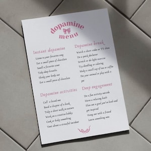 Puede incluir: Un papel blanco titulado "menú de dopamina" con texto rosa y una ilustración de lazo. El menú enumera actividades para la dopamina instantánea, descansos de dopamina, actividades de dopamina y compromiso profundo.