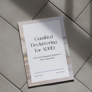 May include: A white and beige book titled "Gamified Decluttering for ADHD: A Fun and Engaging System to Stay Organized." The book is on a light gray tiled surface. The author's name and the company name are at the bottom.