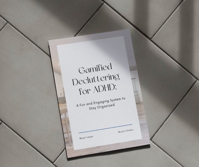 May include: A white and beige book titled "Gamified Decluttering for ADHD: A Fun and Engaging System to Stay Organized." The book is on a light gray tiled surface. The author's name and the company name are at the bottom.