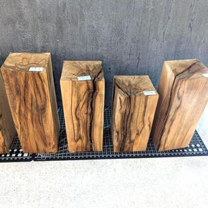 Könnte beinhalten: Fünf rechteckige Holzblöcke mit natürlichen Holzmaserungsmustern. Die Blöcke variieren in der Größe und sind auf einem schwarz-weißen Raster angeordnet. Das Holz hat eine warme, hellbraune Farbe mit dunkleren braunen Streifen.