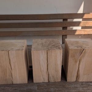 Könnte beinhalten: Drei rechteckige Blöcke aus hellem Naturholz, die nebeneinander angeordnet sind. Jeder Block weist eine sichtbare vertikale Maserung und einen deutlichen natürlichen Riss in der Mitte der Vorderseite auf, was die rohe Textur unterstreicht.