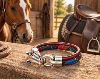Op de afbeelding: Een armband bestaande uit een donkerrood leren koord gecombineerd met een veelkleurige geweven stoffen band. Het sieraad is voorzien van een zilverkleurige metalen sluiting in de vorm van een paardenkop, verbonden met een ring en eindkap.