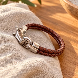 May include: A brown braided leather bracelet featuring a silver-toned metal clasp shaped like a horse head. The bracelet consists of two strands of woven leather that connect to the metallic horse head ornament and a matching silver-toned link.