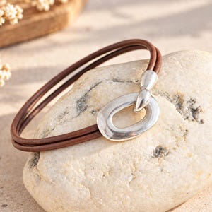 Könnte beinhalten: Ein Armband aus zwei braunen Lederbändern mit einem ovalen Verschluss aus silberfarbenem Metall. Das Armband besteht aus zwei dünnen Lederkordeln, die an einem glatten, abgerundeten, ovalen Glied als Verschlussmechanismus befestigt sind.