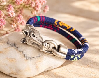 Op de afbeelding: Een geweven stoffen armband met een donkerblauwe basis en veelkleurige geometrische patronen in geel, groen en wit. De armband is voorzien van een zilverkleurige metalen sluiting in de vorm van een paardenkop en een bijpassende lus.